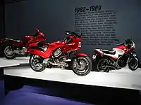 Three sporty motorcycles of the 1990s on pedestals. Text on the wall behind them says "1982-1989" with a few dozen words relevant to the period, such as "Atari", "MTV", and "Robert Mapplethorpe".