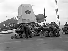 Black and white photograph of a group of men pushing bombs on trolleys on the deck of an aircraft carrier at sea. A single-engined aircraft is located immediately behind the men.