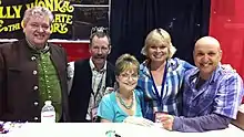 Five middle-aged white adults (two women and three men) in casual dress are behind a table, posing for a group photo.