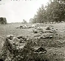 Square black-and-white photograph showing bodies on the ground. A wagon can be seen on the left.