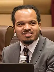 A young, black man with a mustache and anchor beard sits behind a laptop and stares off to his left. Another person, barely visible, is seated to Ellison's right.
