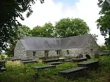 A T-shaped low stone church with a slate roof seen beyond grave slabs
