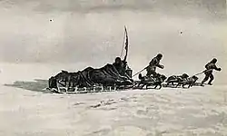 A loaded sledge being pulled across an icy surface by two figures and a team of dogs