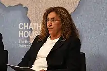 Middle Eastern woman with long hair, wearing glasses, a white top and dark jacket, sitting and looking ahead.