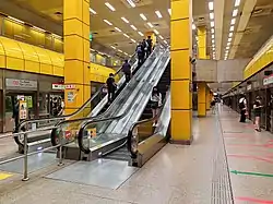 Underground island platform with escalators between two yellow pillars leading up to the concourse