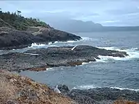 A rock outcropping in a bay being hit by waves of water with a mountain in the far distance
